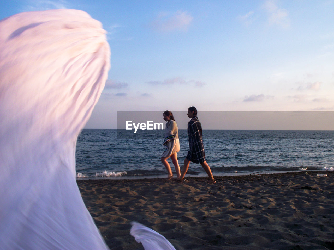 Friends with towels walking at beach against sky during sunset