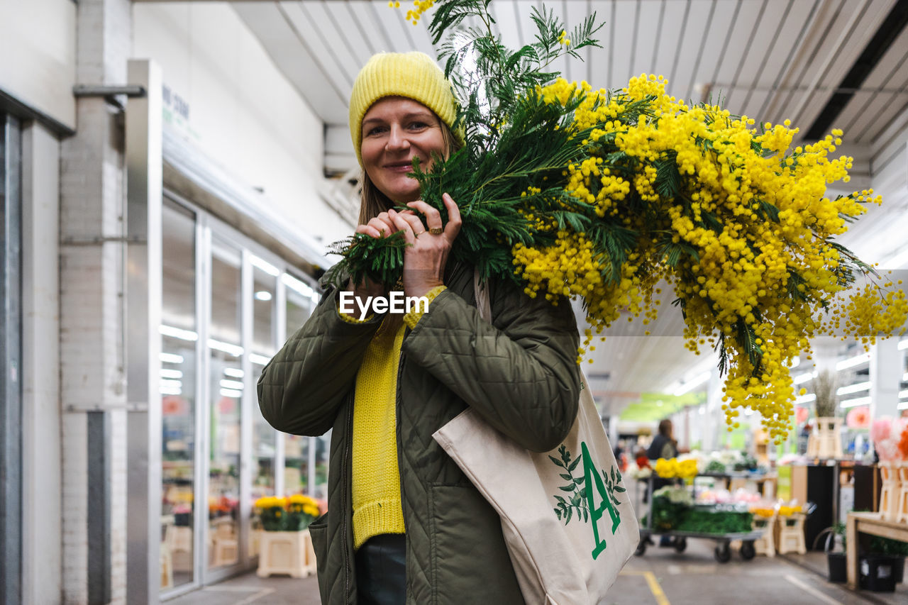 Woman in yellow cap and large bouquet of mimosa flowers at flower market