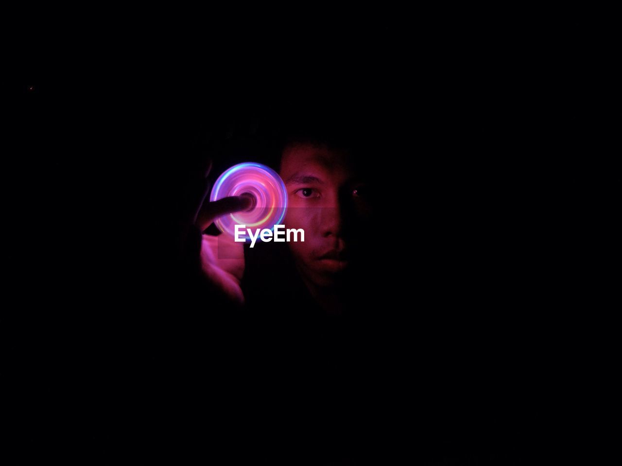 Portrait of young man spinning illuminated fidget spinner against black background