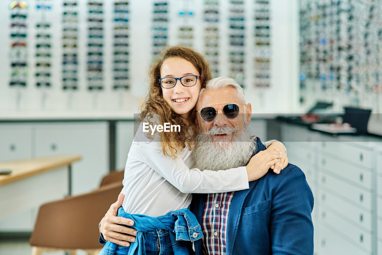 Smiling stylish grandpa and cute girl wearing trendy glasses while embracing in modern optical store and looking at camera