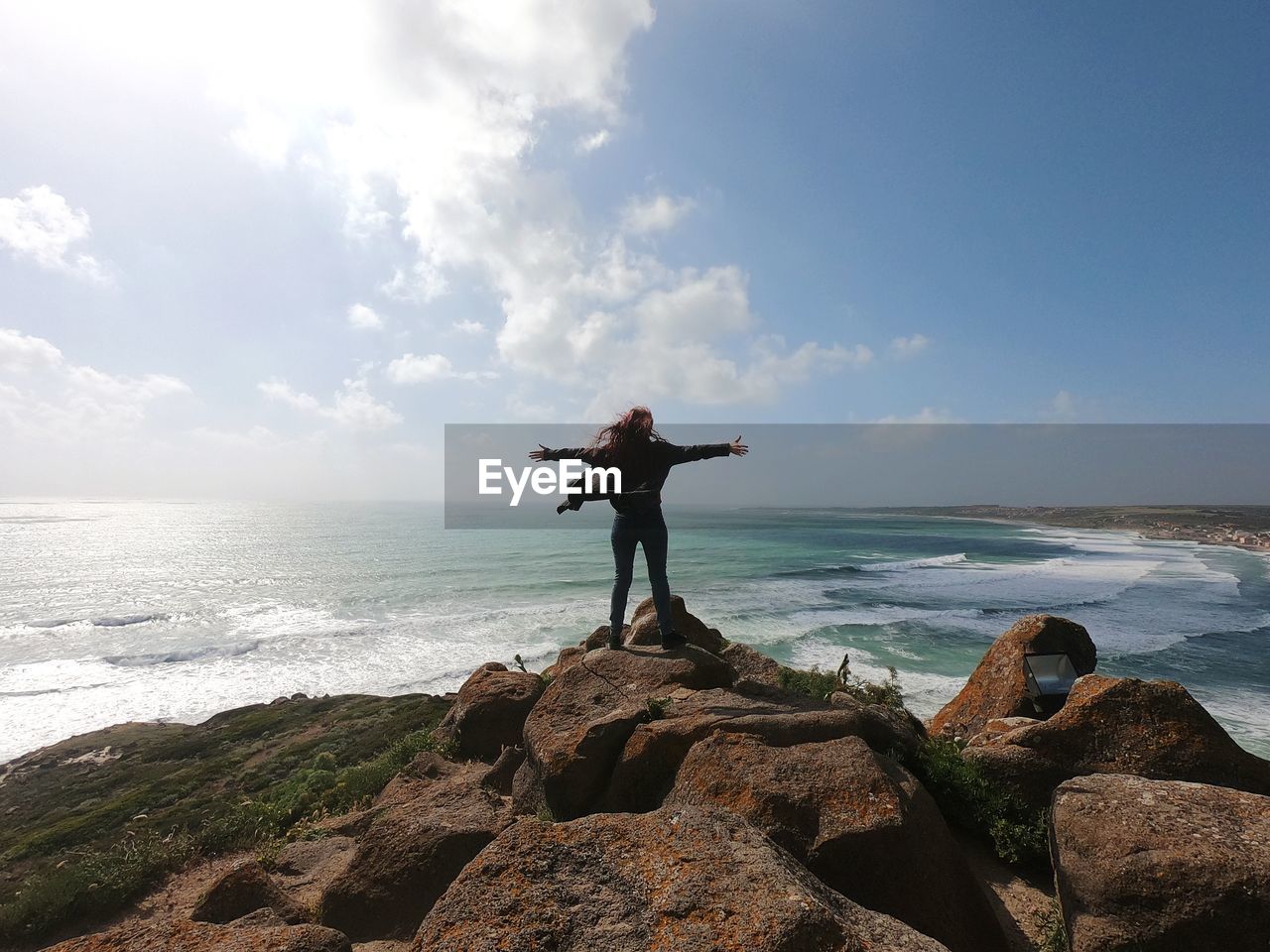 Rear view of woman with arms outstretched standing on rock at beach against cloudy sky