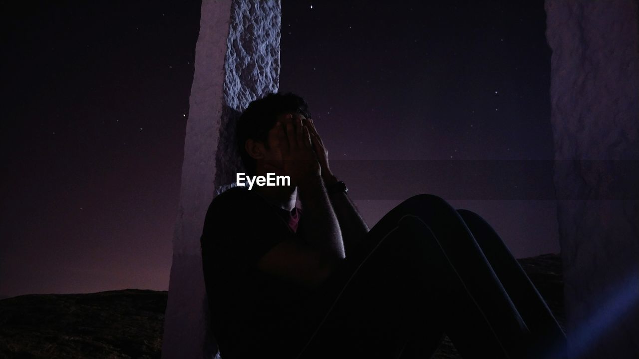 Man with face covered by hands sitting against sky at night