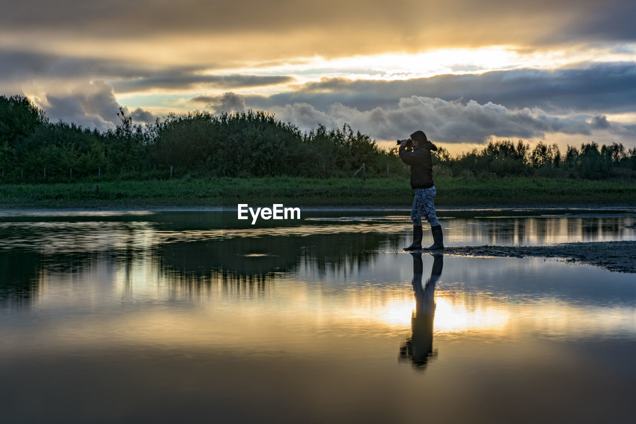 Man photographing at lakeshore against cloudy sky during sunset