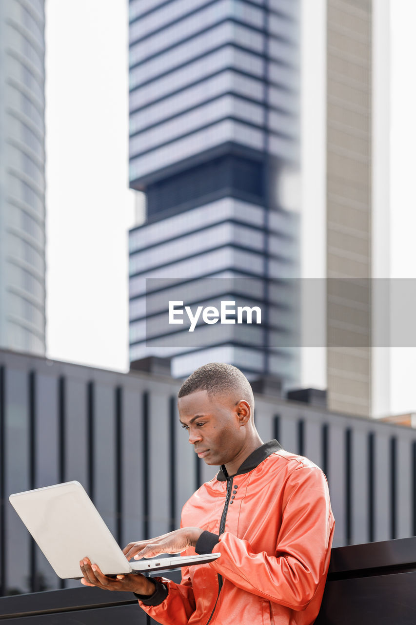 Side view of focused young african american male student in stylish outfit and backpack standing on street and using computer while preparing for exams on sunny day