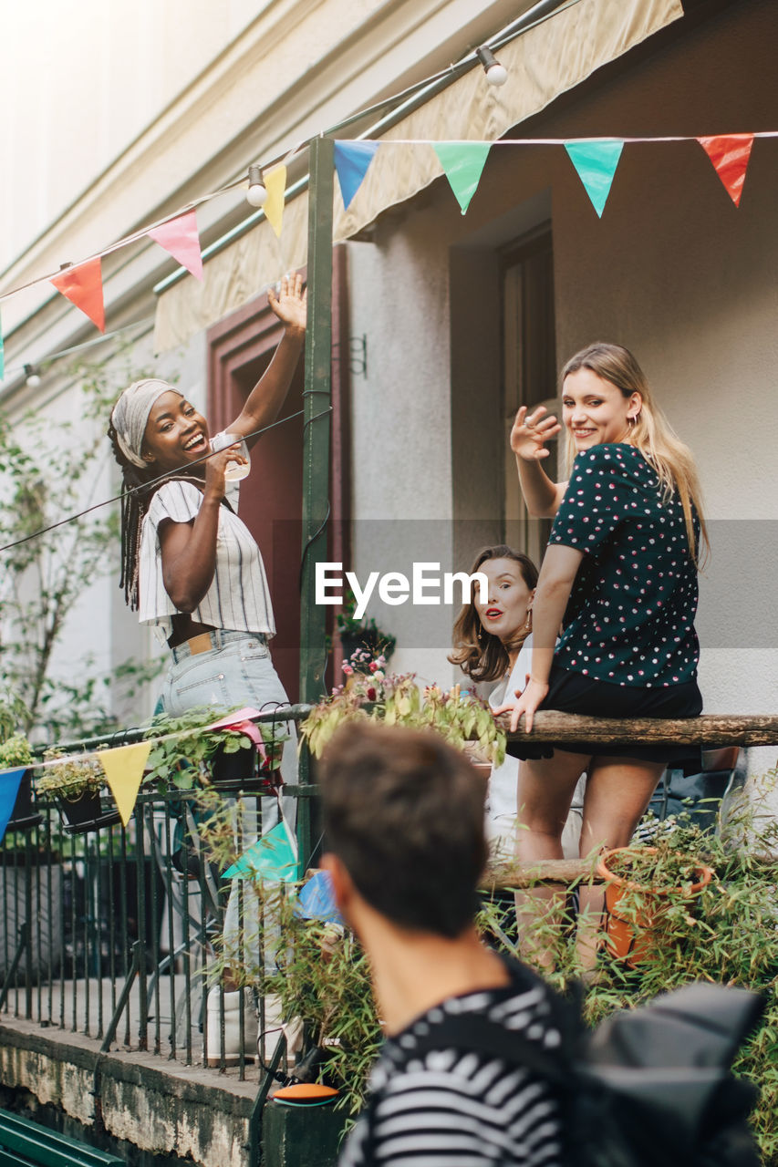 Happy young women waving hands at male friend while standing in balcony during party