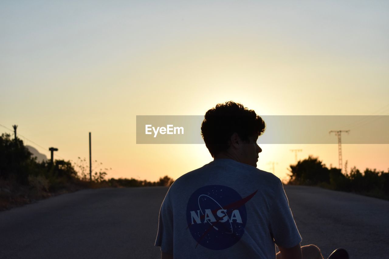 REAR VIEW OF MAN STANDING ON ROAD AT SUNSET