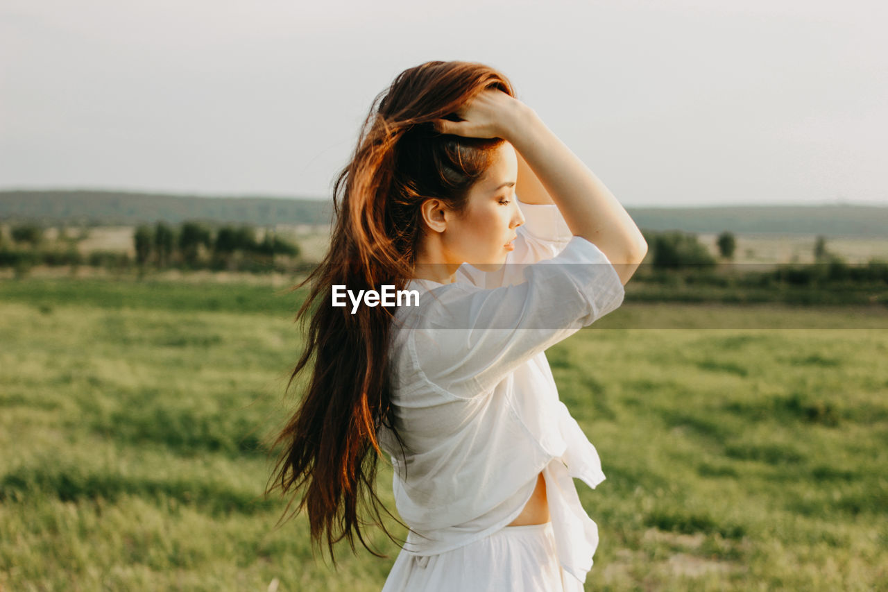 Beautiful young woman with hands in hair standing on land against sky