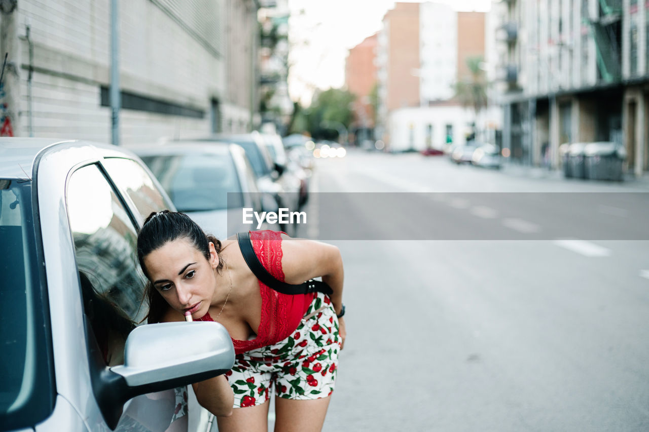 Stylish young woman standing near car on roadside and looking at side mirror while applying lipstick on city street