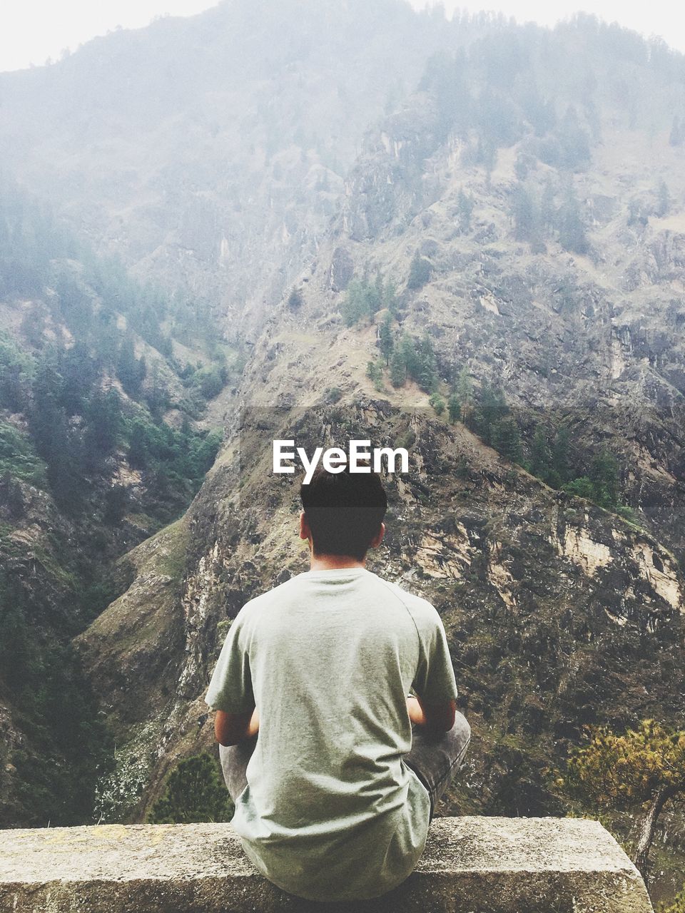 Rear view of man looking at mountains while sitting on concrete wall