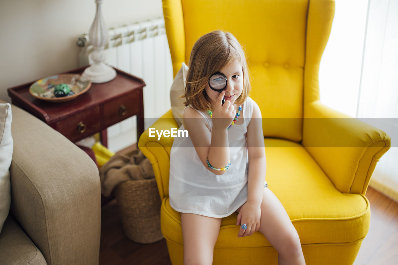 Blonde little girl with a magnifying glass in her eye sitting on a yellow armchair