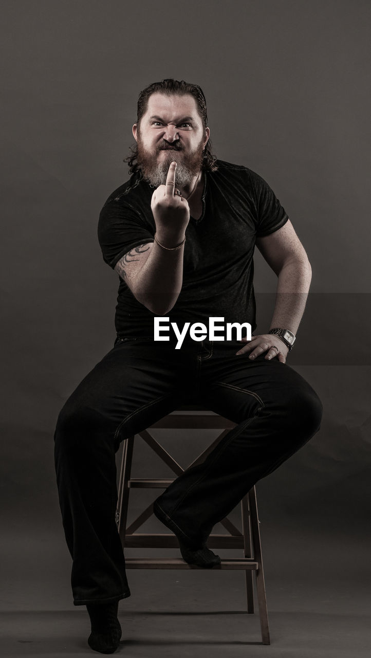 Mid adult man gesturing obscene gesture while sitting against black background