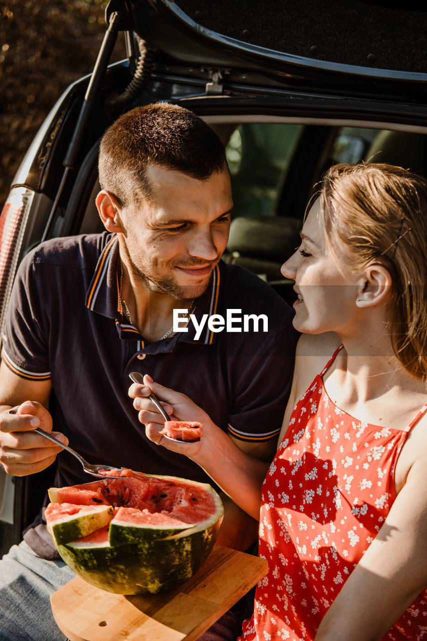 Millennial couple sitting on open trunk and eating watermelon. happy young couple having break at