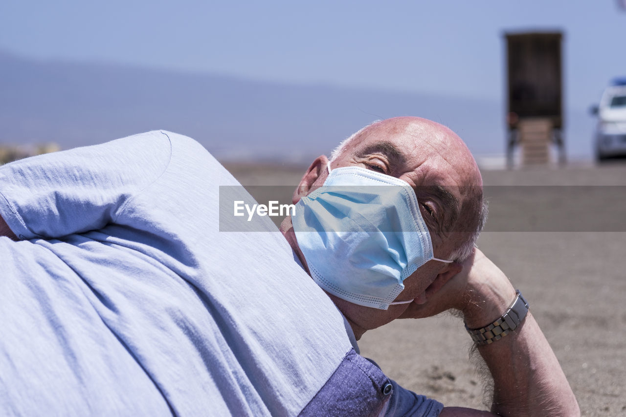 Portrait of senior man wearing flu mask relaxing on beach against sky