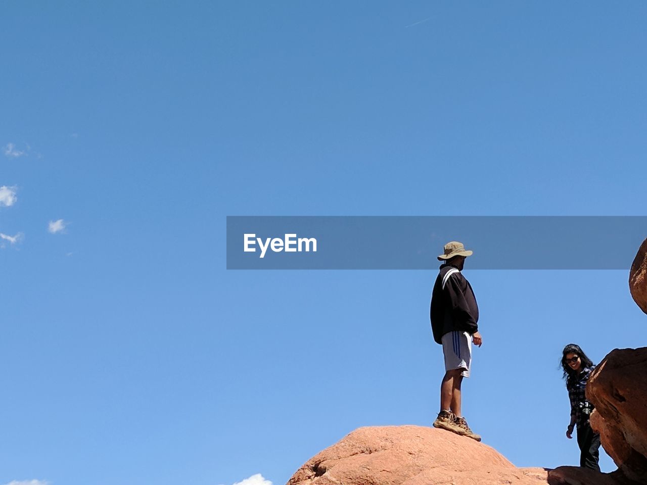 LOW ANGLE VIEW OF PEOPLE ON ROCKS AGAINST CLEAR BLUE SKY