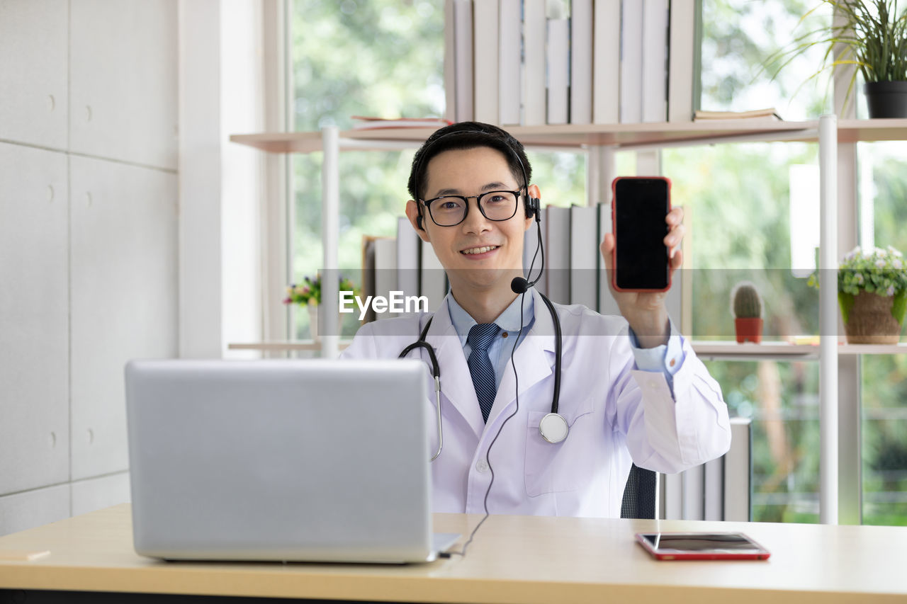 Portrait of smiling doctor showing mobile phone while sitting at hospital