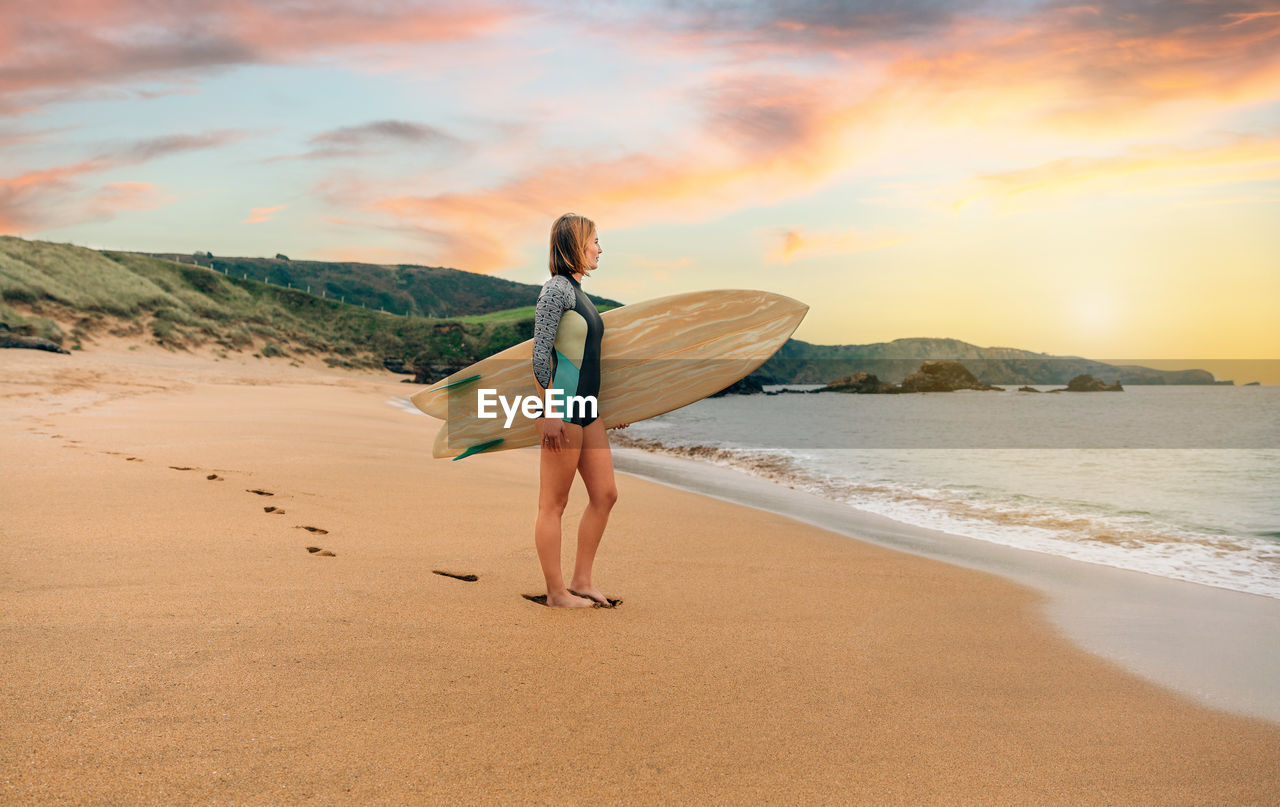 Surfer woman with wetsuit carrying surfboard looking to the sea at the beach