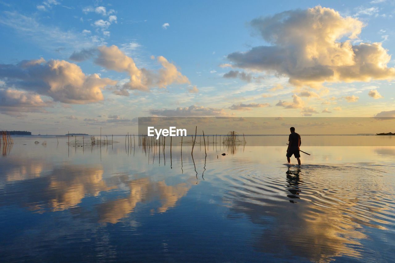 Man walking in sea against sky during sunset