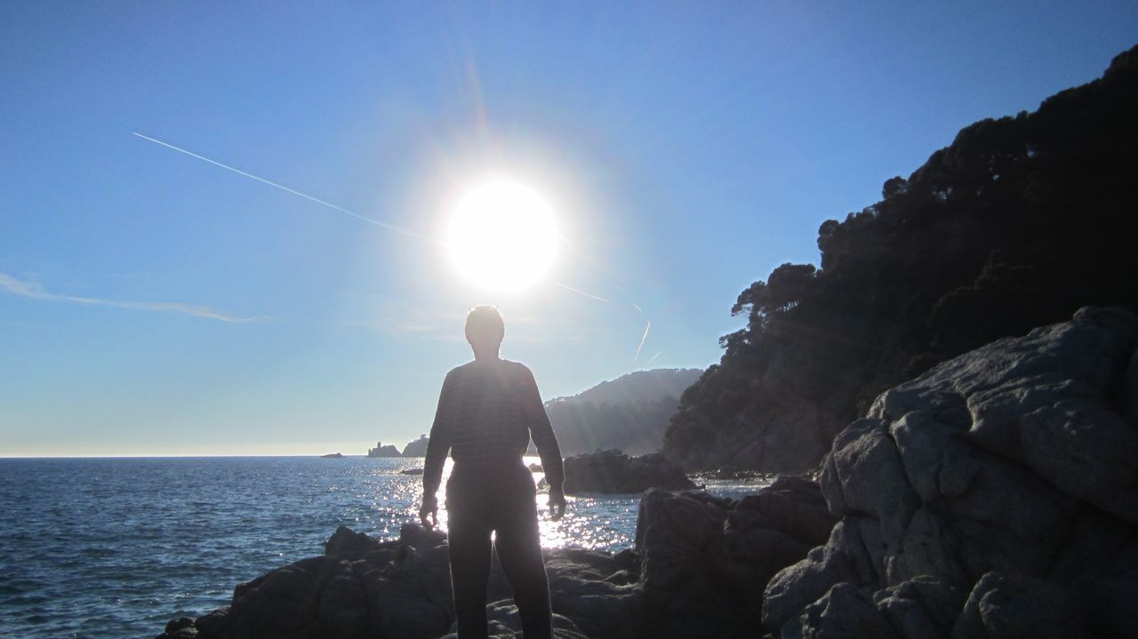 Rear view of silhouette man standing on rock by sea against clear sky