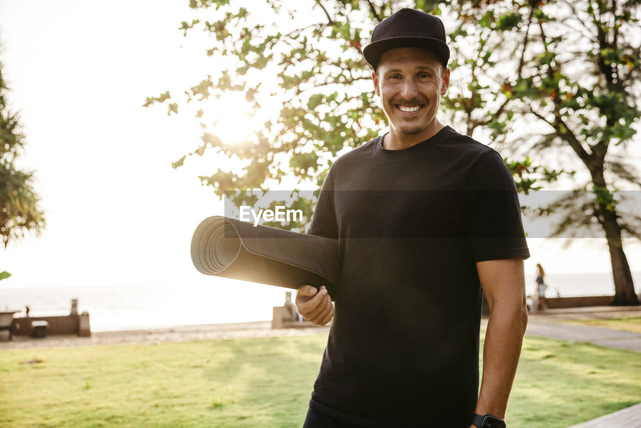 Portrait of smiling man with exercise mat standing in park