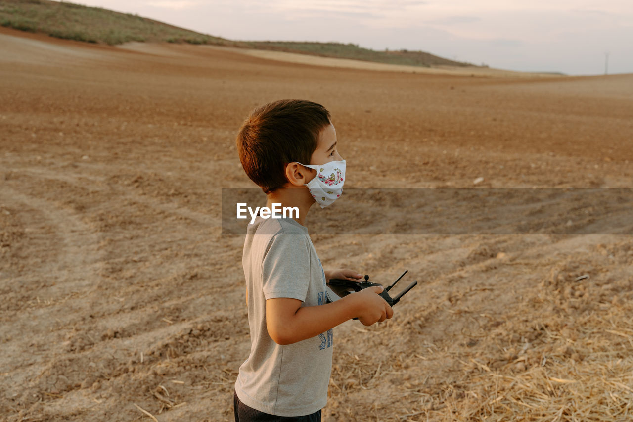 Side view of focused little asian boy in protective mask using remote control while playing with flying drone in field