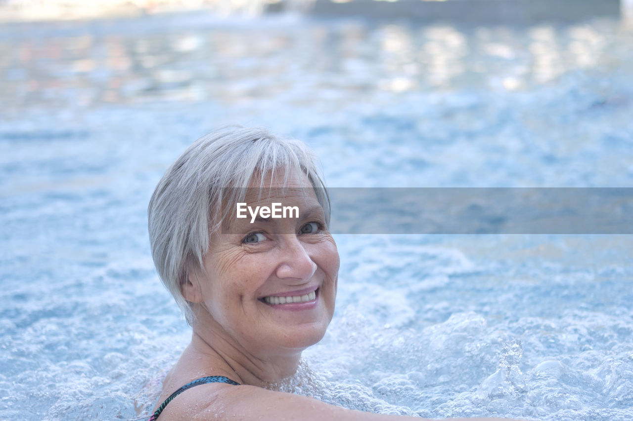 Smiling senior woman with gray hair looking at camera in outdoor thermal pool with hydromassage. 