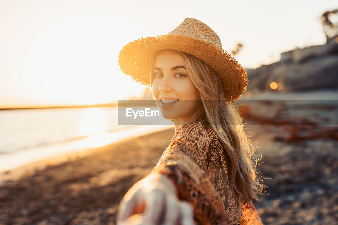 portrait of young woman wearing hat standing at beach against sky during sunset