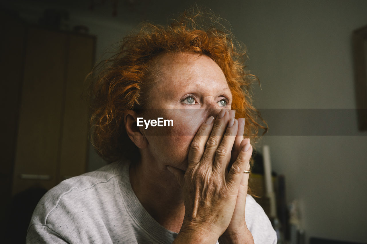 Thoughtful sad senior woman with hands covering mouth sitting at home