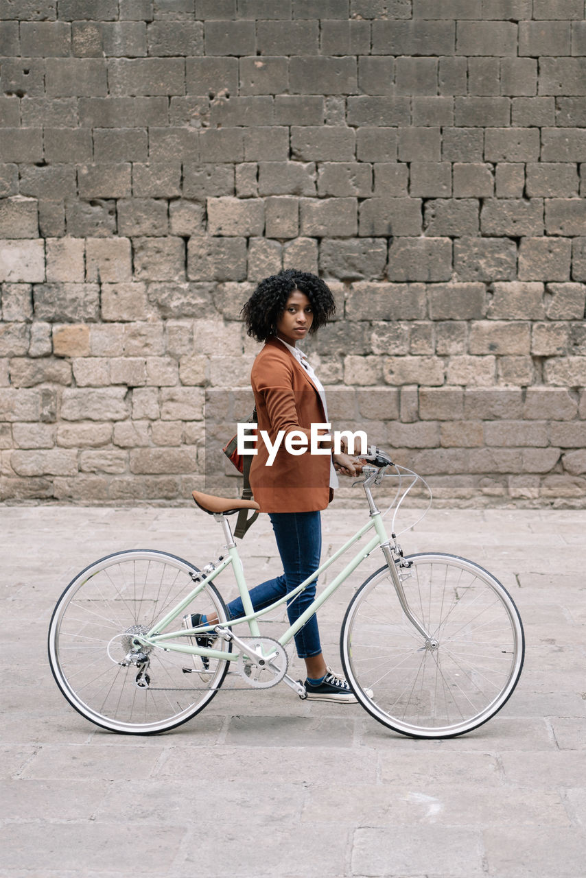 Side view of black woman in smart casual style walking along street with bike and looking at camera