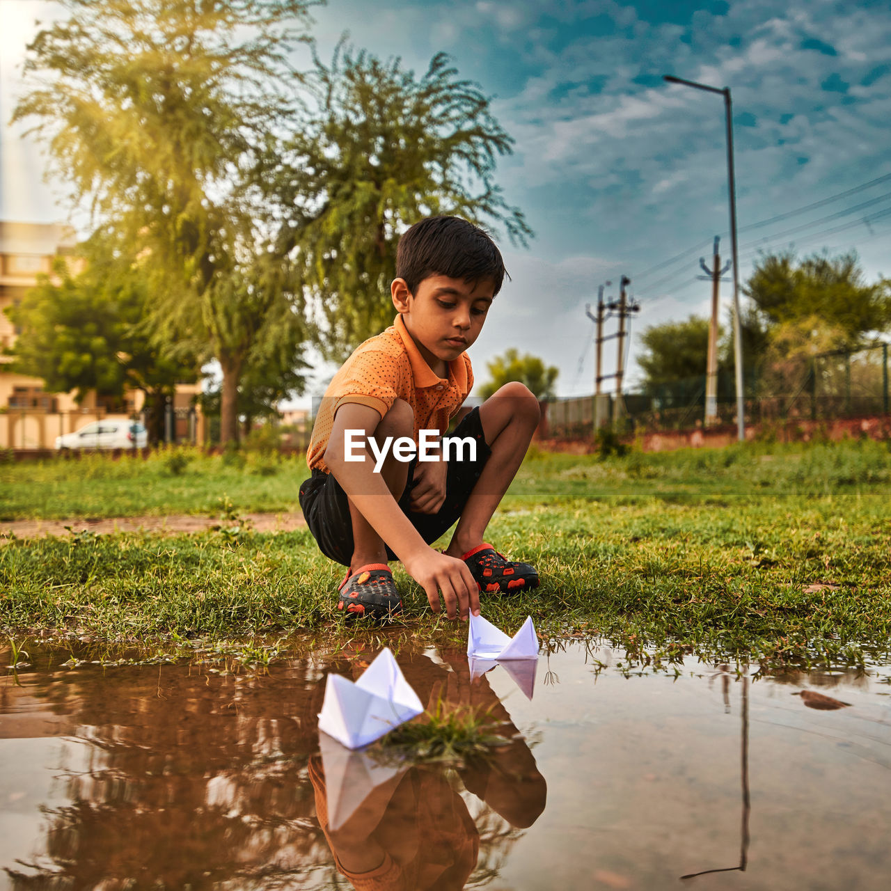 Full length of boy crouching on grass by paper boat in puddle against sky