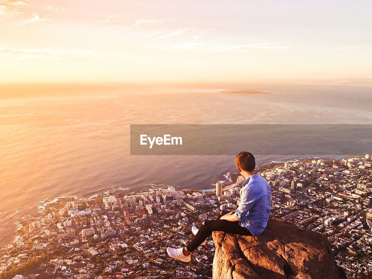 Side view of man sitting on rock over cityscape and seascape during sunset