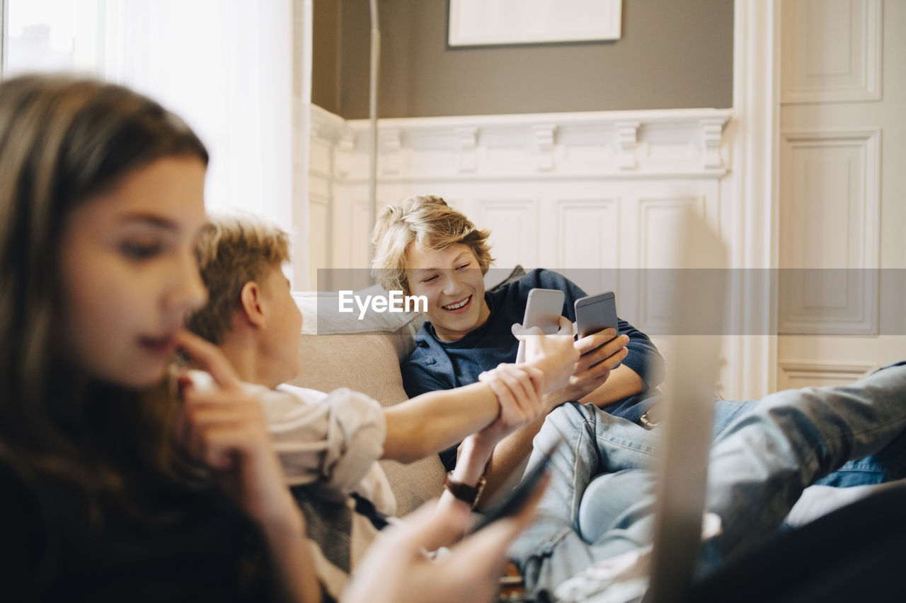 Boys using mobile phone while sitting with female friend on sofa