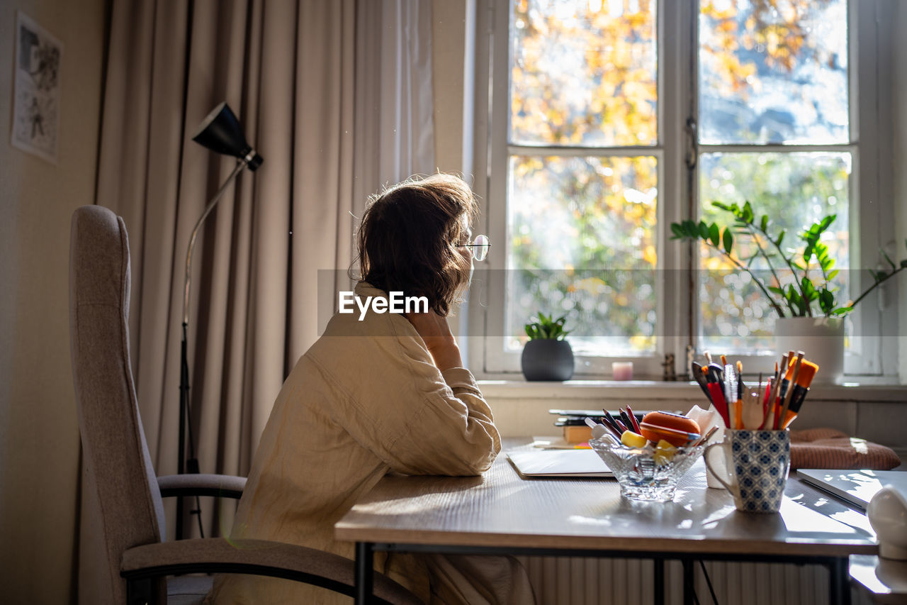 Woman painter having inspiration to create new picture sitting at desk thinking looking at window.