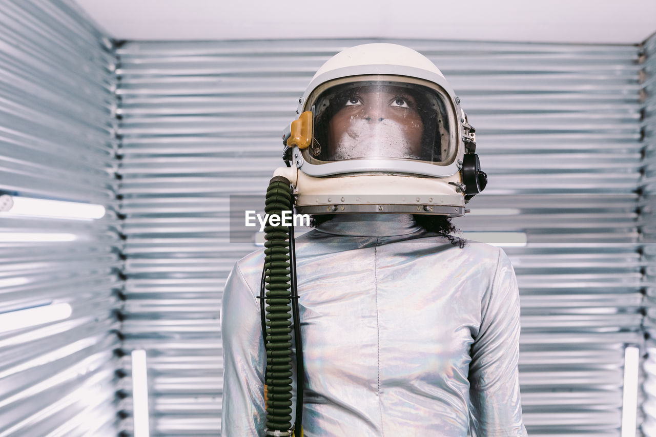 Serious professional african american female astronaut in helmet standing in modern light spaceship with silver walls before going on mission looking up