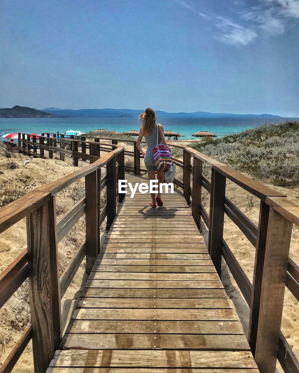 walkway, boardwalk, water, sea, sky, beach, vacation, nature, women, land, full length, wood, leisure activity, one person, rear view, lifestyles, railing, adult, shore, coast, pier, horizon, horizon over water, beauty in nature, day, holiday, sunlight, trip, scenics - nature, female, architecture, casual clothing, body of water, walking, outdoors, the way forward, tranquility, relaxation, sand, footpath, travel, travel destinations, built structure, ocean, tranquil scene, summer, child, sunny, motion, shadow, clothing, blue