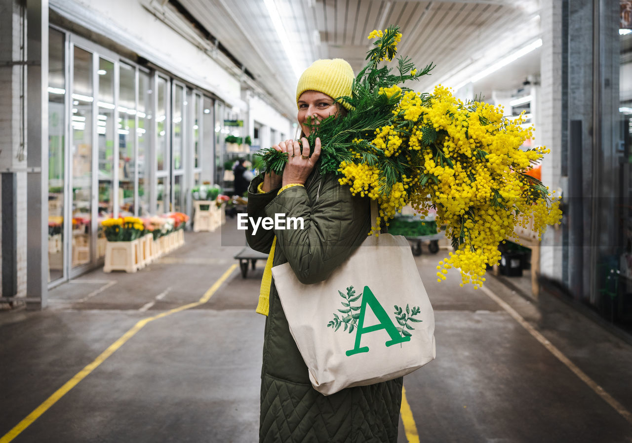 Positive woman in yellow cap and large bouquet of mimosa flowers at flower market