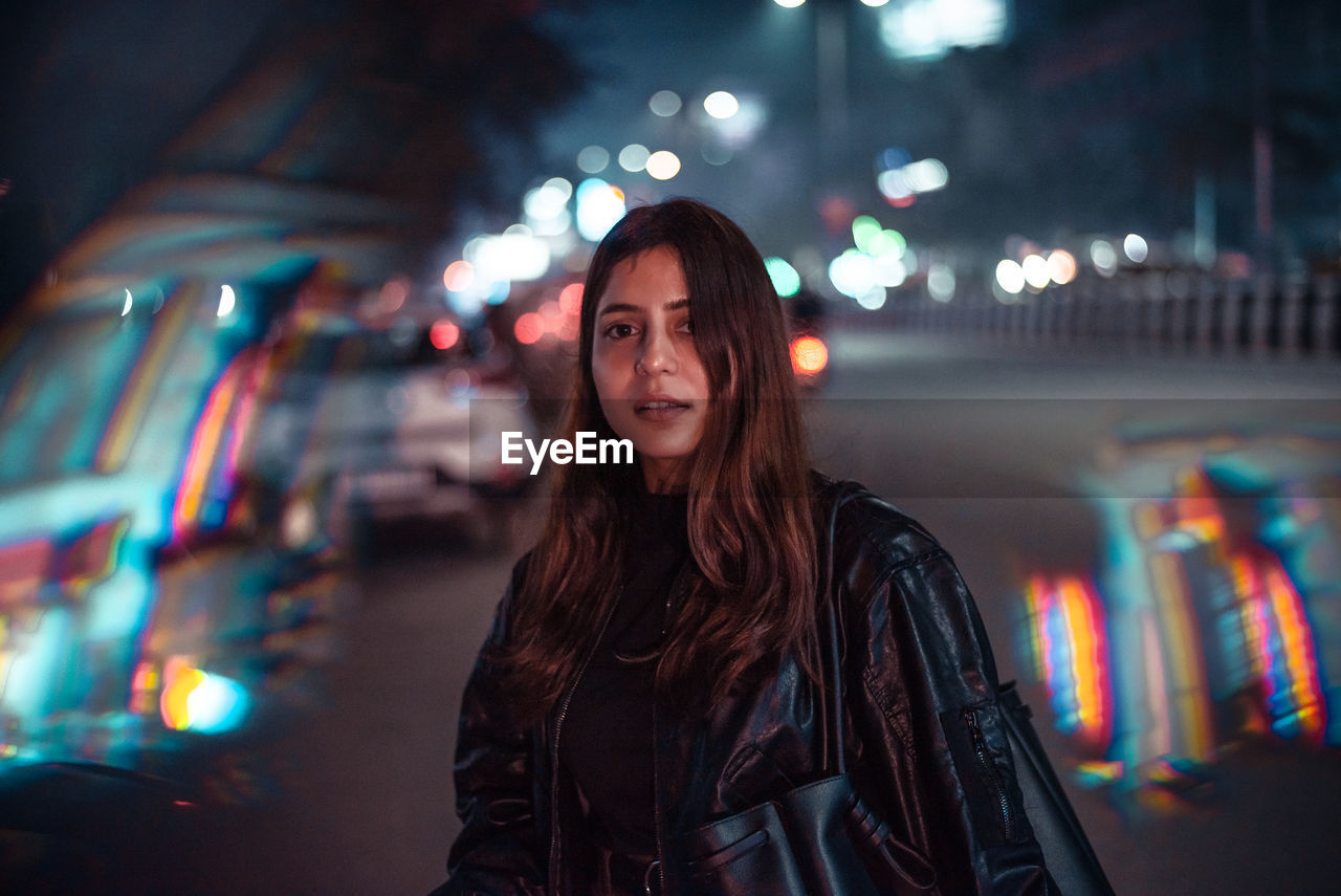 PORTRAIT OF YOUNG WOMAN STANDING AGAINST ILLUMINATED BUILDING