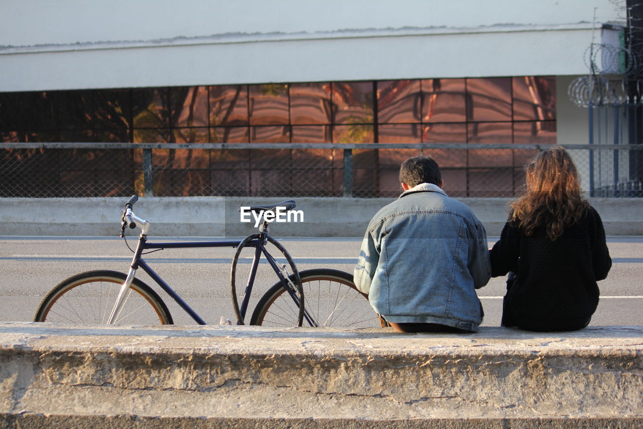 Rear view of couple sitting on retaining wall by bicycle