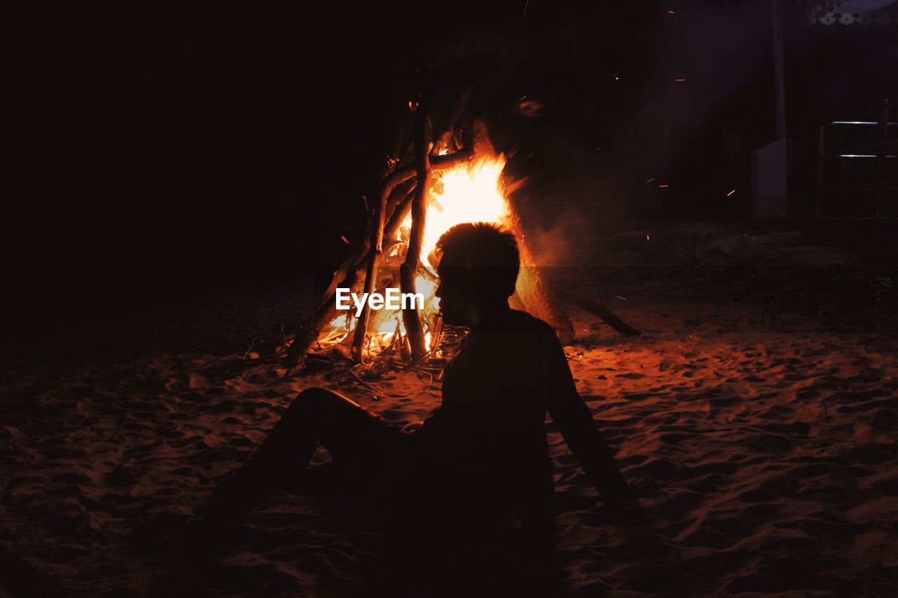 Silhouette man sitting by burning bonfire at campsite