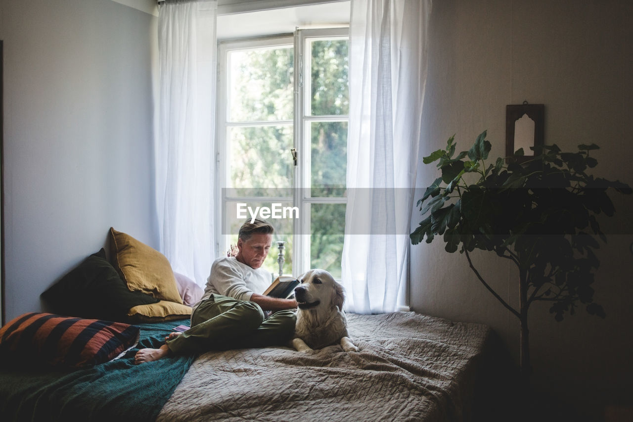 Full length of retired man reading book while resting with dog on bed at home