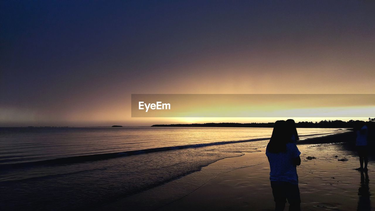 Rear view of silhouette woman walking at beach against sky during sunset