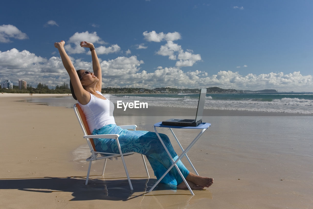 Side view of mid adult woman with arms raised with laptop sitting at beach against sky