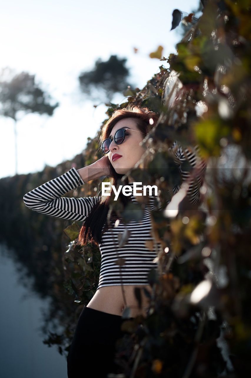 Young woman standing by trees against sky