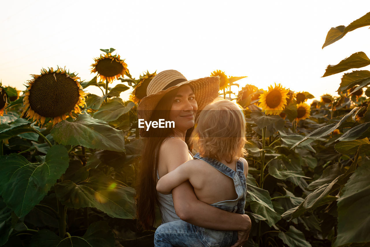 Beautiful mother holds a little son in her arms in a field of sunflowers at sunset