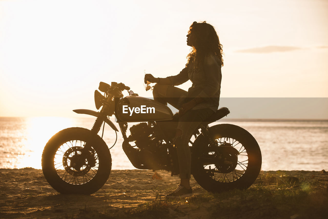 Woman sitting on motorcycle at beach during sunset