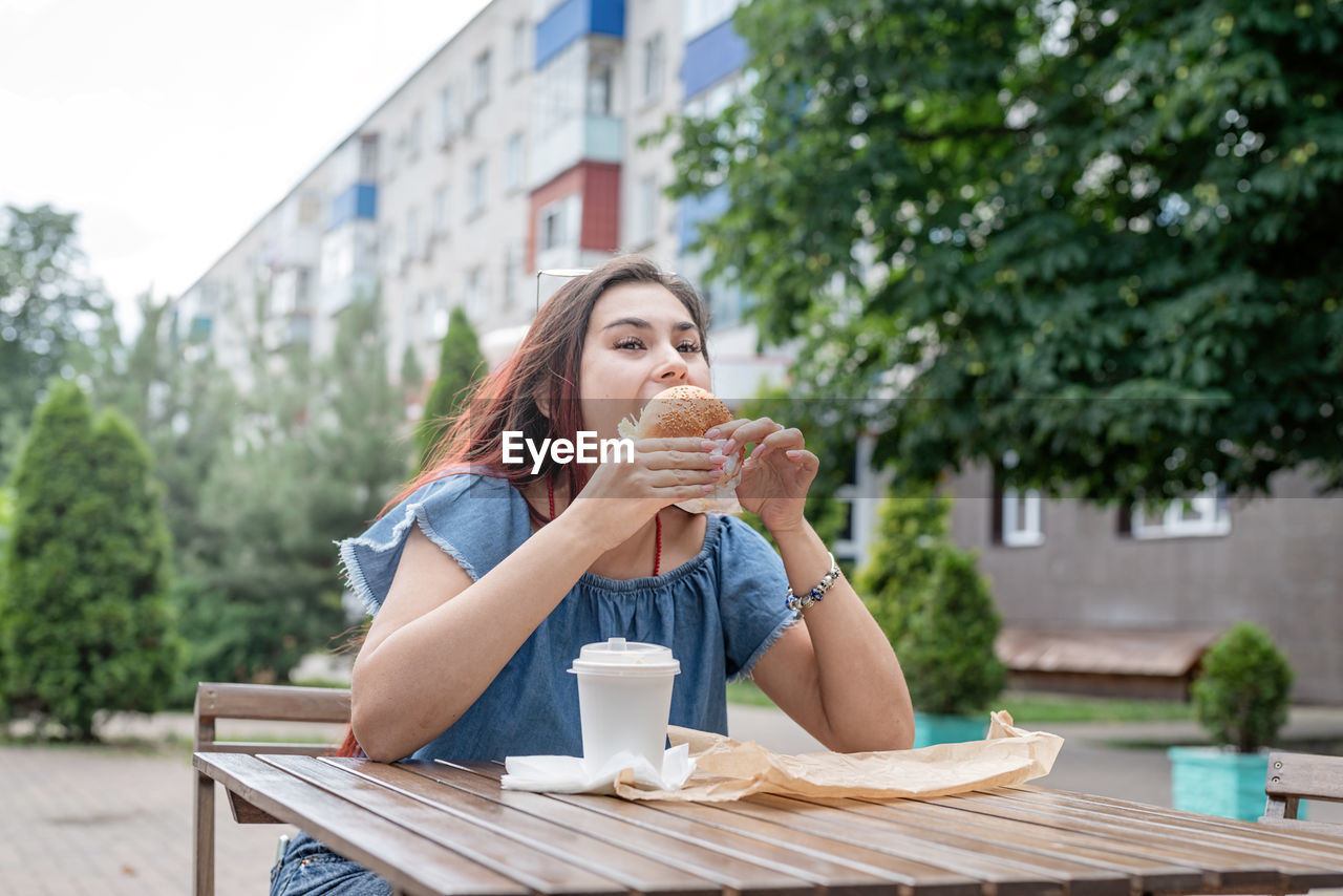 Hungry stylish woman, enjoying eating a burger outdoors, dressed in jeans shirt, wearing sunglasses