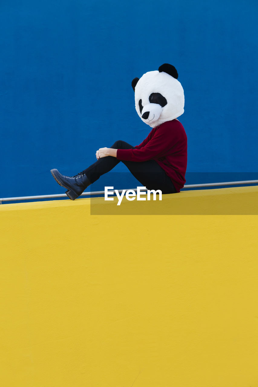 Female wearing panda mask while sitting against blue wall