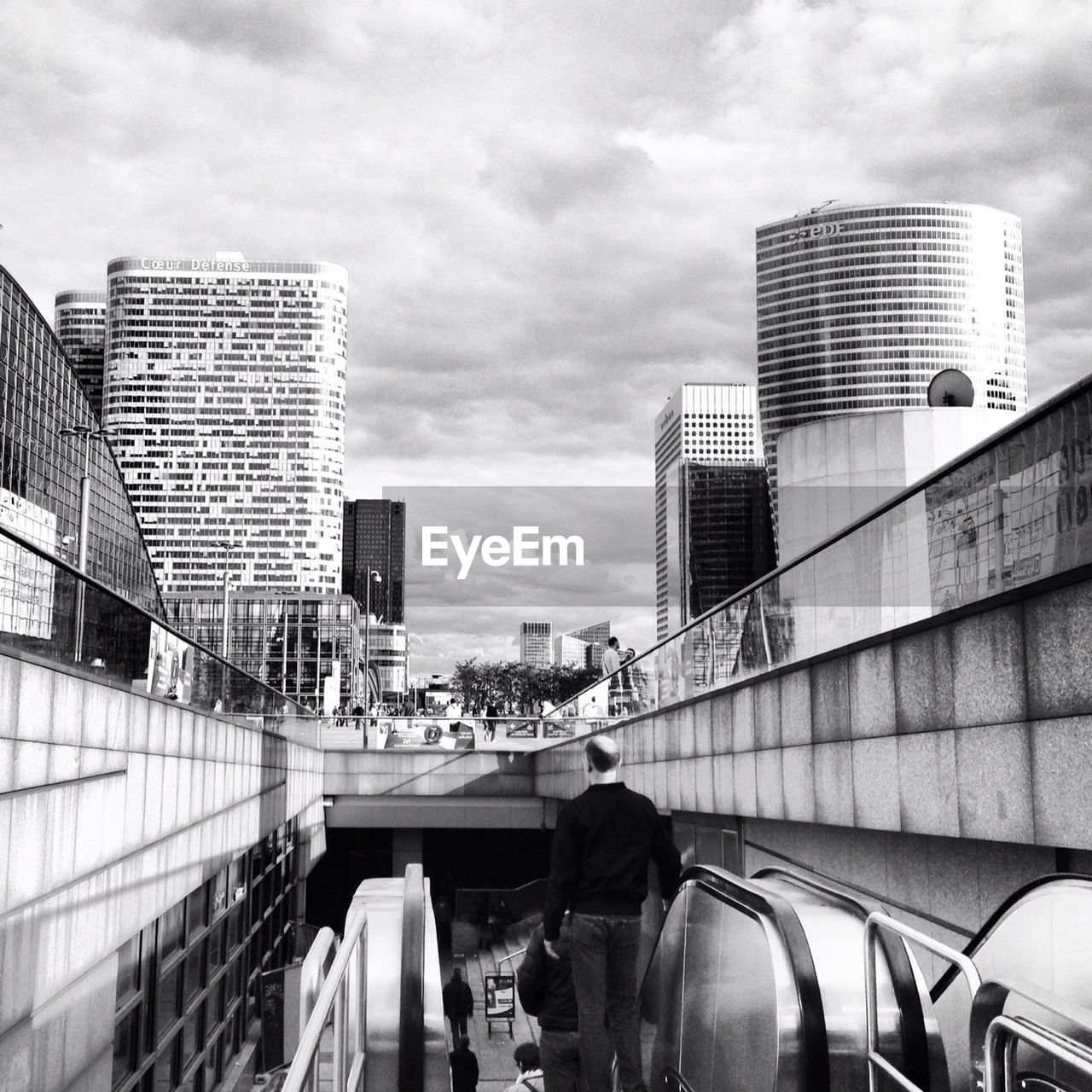 Man on escalator leading towards railroad station in city against cloudy sky