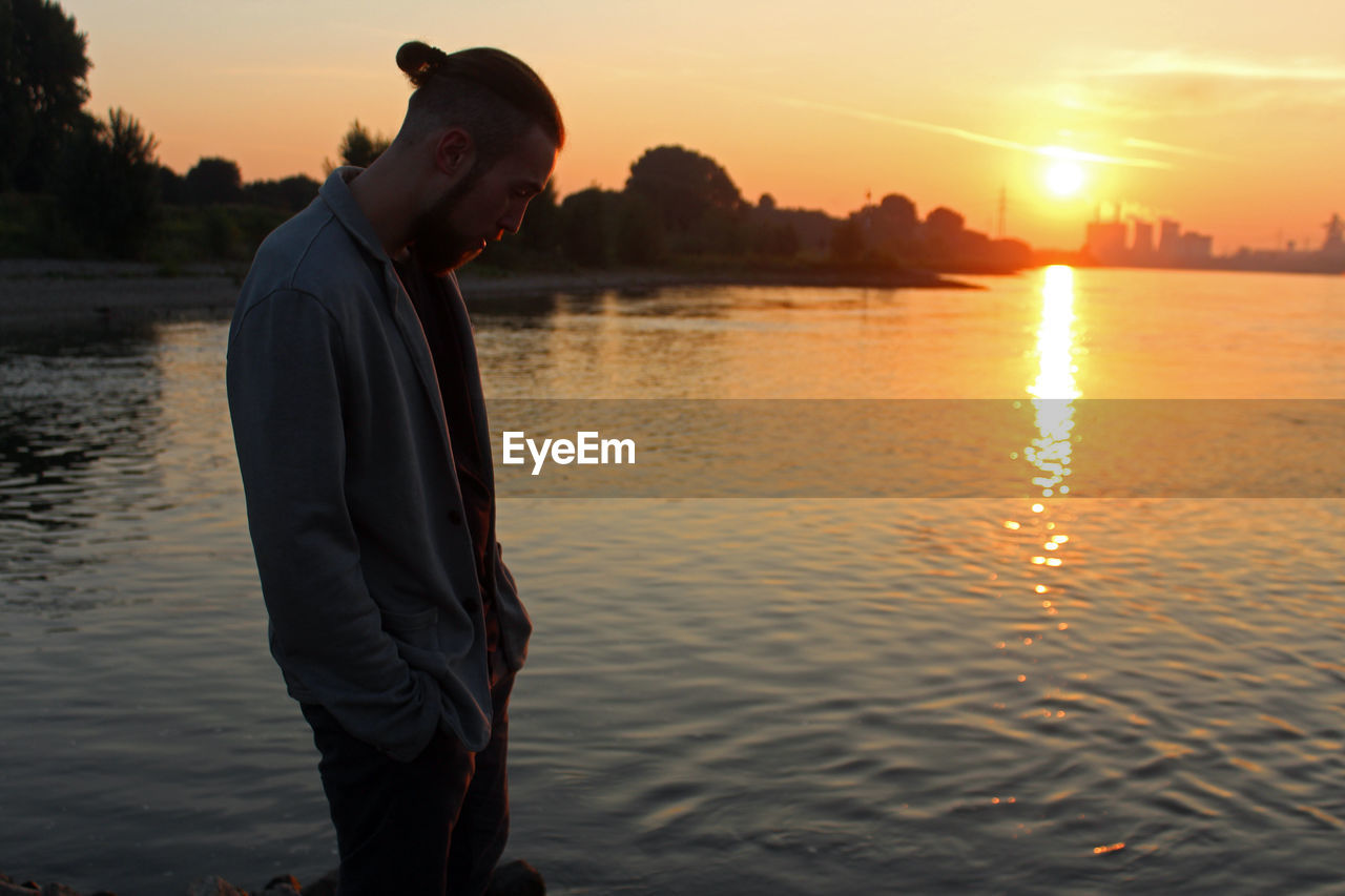 Side view of young man standing by lake against sky during sunset