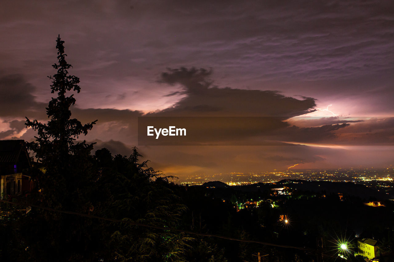 SILHOUETTE TREES AND ILLUMINATED CITY AGAINST DRAMATIC SKY