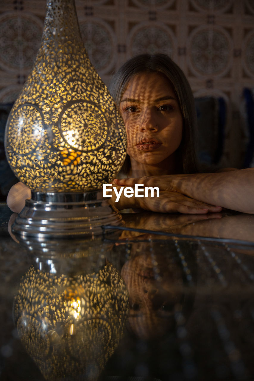 Portrait of young woman sitting against illuminated lamp at home